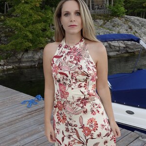 May include: A cream halter-neck dress featuring a floral design in red and pink hues. The dress has a fitted top and a flowing skirt. The model is standing on a wooden pier near water, with a boat visible in the background.