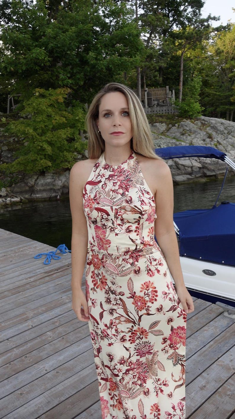 May include: A cream halter-neck dress featuring a floral design in red and pink hues. The dress has a fitted top and a flowing skirt. The model is standing on a wooden pier near water, with a boat visible in the background.