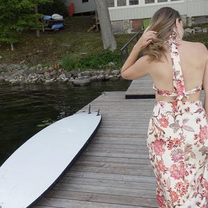 May include: A white paddleboard rests on a wooden dock next to a woman wearing a floral print halter top and skirt. The outfit features a cream background with pink and red floral patterns. The dock extends over a lake.