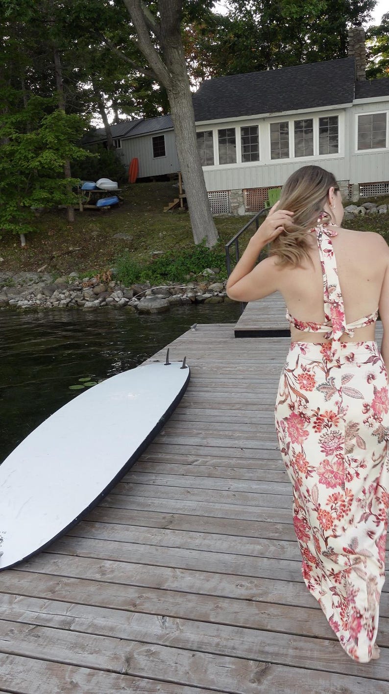 May include: A white paddleboard rests on a wooden dock next to a woman wearing a floral print halter top and skirt. The outfit features a cream background with pink and red floral patterns. The dock extends over a lake.