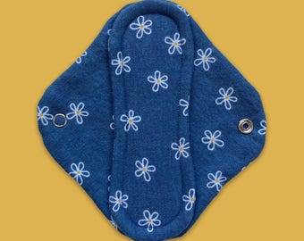 May include: A reusable, blue cloth menstrual pad with a white floral pattern. The pad has a contoured shape with snaps for secure closure. The background is a solid mustard yellow.