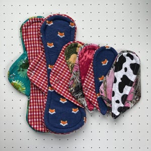 May include: A collection of reusable cloth menstrual pads in various patterns including a dark blue fox print, red and white gingham, green with frog illustrations, pink camouflage, and a black and white cow print, each featuring a metal snap closure.