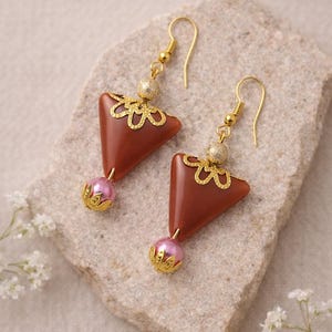 Op de afbeelding: Oorbellen met een driehoekig, roodbruin ontwerp, geaccentueerd met gouden filigraan en een roze parel. De oorbellen hangen aan gouden haken, tegen een neutrale stenen achtergrond.