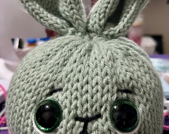 Könnte beinhalten: Ein handgestrickter grauer Hase mit grünen Augen und einem weißen Gesicht. Der Hase ist aus weichem Garn gefertigt und hat ein süßes, kuscheliges Aussehen.