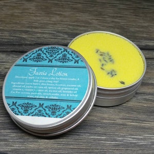 May include: A small, open, silver-colored tin of yellow lotion with lavender buds, next to a closed tin with a teal label. The label reads "Faerie Lotion" with a list of ingredients and uses.