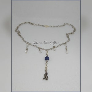 Op de afbeelding: Een zilverkleurige ketting met een fijne ketting. De ketting heeft heldere kristallen accenten, een donkerblauwe kraal en een kleine hanger van een zittende figuur. De tekst "Faerie Ever After" is zichtbaar.