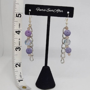 May include: Dangle earrings with lavender lava beads, iridescent crystal beads, and silver-tone chain links. The earrings are displayed on a black stand. The text "Faerie Ever After" is above the earrings. The earrings are approximately 3 inches long.