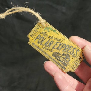 May include: A rectangular, glittery yellow tag with the words "POLAR EXPRESS" and "BELIEVE" printed in black. The tag has a train illustration and is attached to a natural twine string.