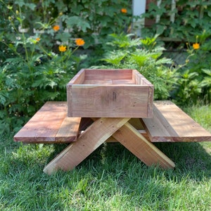 May include: A small wooden picnic table with a built-in planter box. The table is made of natural cedar wood and has a rustic finish. The planter box is located in the centre of the table and is perfect for growing small plants or herbs.