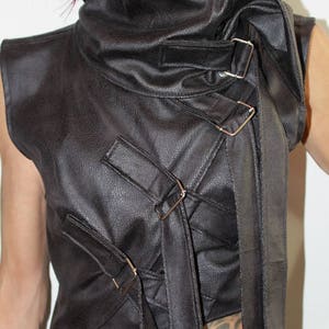 Pode incluir: Um colete preto de couro falso com gola alta e várias tiras e fivelas. O colete tem um comprimento curto e é projetado para ser usado aberto.