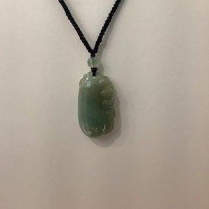May include: A green jade pendant carved with a floral design, hanging from a black cord necklace.