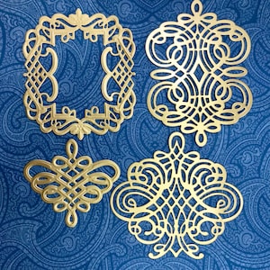 May include: Four gold-colored metal filigree embellishments with intricate, symmetrical, and openwork scroll patterns. The collection includes one rectangular frame and three distinct decorative shapes featuring interconnected loops and curves.
