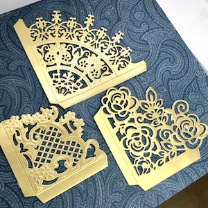 May include: Three gold-colored metal corner protectors with intricate cut-out designs. One piece features a fan-like pattern, another displays a grid and floral motif, and the third showcases a detailed rose and leaf pattern with smooth edges.