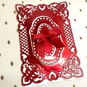 May include: A rectangular card featuring a shiny red metallic die-cut frame with intricate heart and lattice patterns. The center displays nested oval layers with heart-shaped cutouts, topped with a tied red satin ribbon bow in the middle.