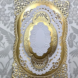 May include: A layered die-cut paper craft piece featuring a rectangular gold metallic base with intricate filigree corners, topped with a white scalloped lace-like frame, a smaller gold oval with textured edges, and a central white oval with embossed roses.