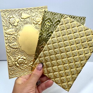 May include: Three rectangular gold-colored sheets with embossed patterns. One sheet features a central blank oval frame surrounded by floral motifs, another displays a repeating fan-like shell design, and the third shows an intricate gothic arch pattern.