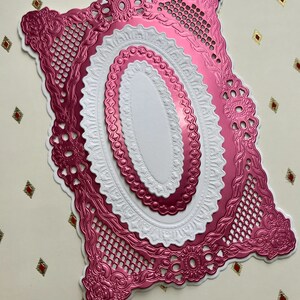May include: A layered die-cut paper craft piece featuring a rectangular metallic pink frame with intricate floral and lattice patterns, surrounding nested white and metallic pink oval shapes with scalloped edges on a cream background.