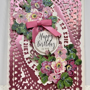 May include: A rectangular greeting card featuring a metallic pink die-cut frame with intricate lattice patterns. The card is decorated with two clusters of glittery pink and white flowers, green leaves, and a pink satin ribbon bow above a circular tag.