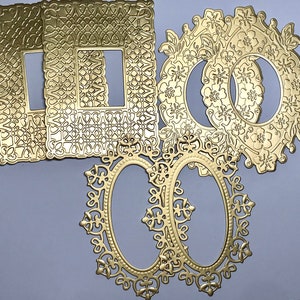 May include: A collection of six gold-colored metal craft dies featuring intricate cut-out designs. Two rectangular frames have geometric lattice patterns, two circular frames feature floral motifs, and two oval frames have ornate filigree borders.