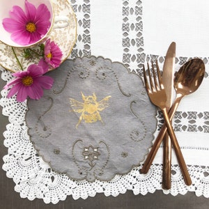 May include: A grey linen doily with an embroidered bee design and delicate lace trim. Three copper-coloured silverware pieces are arranged on a white crocheted tablecloth.