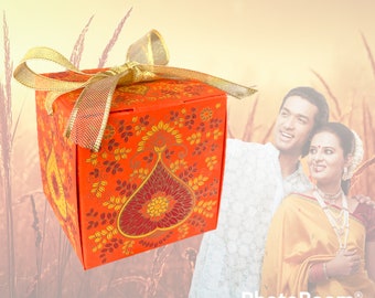 Puede incluir: Una pequeña caja de regalo naranja con una cinta dorada y un patrón floral. La caja está atada con un lazo y tiene un diseño decorativo.