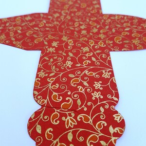 May include: Red and gold patterned paper favor box with a scalloped edge. The box is shaped like a cross with a rounded top.