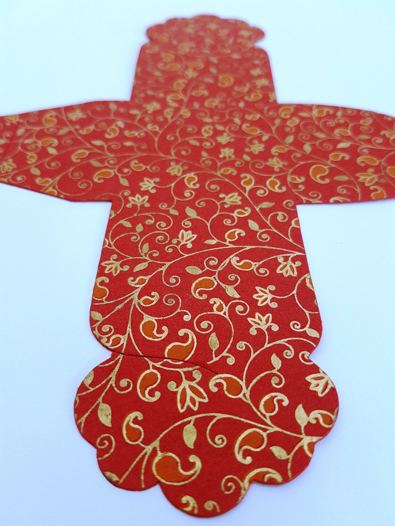 May include: Red and gold patterned paper favor box with a scalloped edge. The box is shaped like a cross with a rounded top.