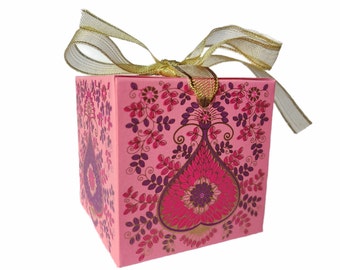 Puede incluir: Una caja de regalo cuadrada rosa con un lazo de cinta dorada. La caja está decorada con un diseño floral en tonos rosa, morado y dorado. La caja tiene forma de cubo.