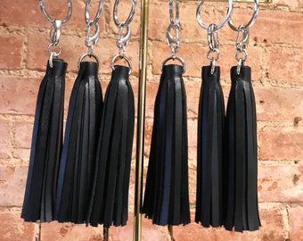 May include: Six black leather tassel keychains hanging on a gold metal stand. The keychains are attached to silver key rings.