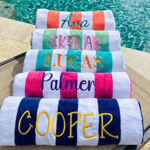 May include: White beach towels with colorful striped borders and embroidered names: Ava, Skylar, Lucas, Palmer, and Cooper. The towels are arranged near a pool, with the water in the background.