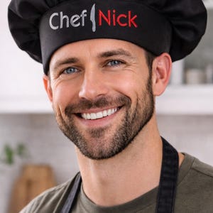 May include: Black chef's hat with the text "Chef Nick" embroidered in silver and red. The hat is worn by a person wearing a black apron and a green shirt. The image is a close-up shot.