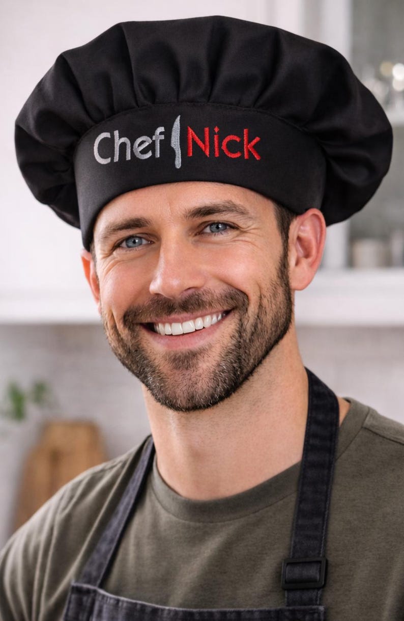 May include: Black chef's hat with the text "Chef Nick" embroidered in silver and red. The hat is worn by a person wearing a black apron and a green shirt. The image is a close-up shot.