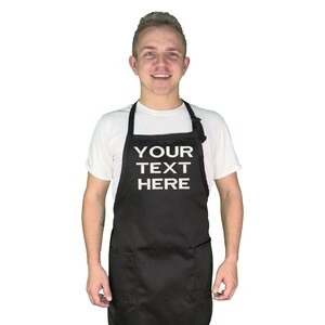 Może przedstawiać: Czarny fartuch z białym napisem "YOUR TEXT HERE". Fartuch ma dwie przednie kieszenie oraz regulowane paski na szyję i w talii. Osoba ma na sobie biały t-shirt.