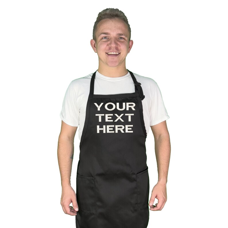 Może przedstawiać: Czarny fartuch z białym napisem "YOUR TEXT HERE". Fartuch ma dwie przednie kieszenie oraz regulowane paski na szyję i w talii. Osoba ma na sobie biały t-shirt.