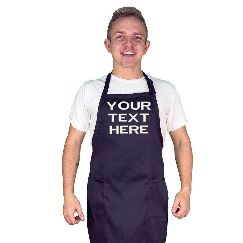 Może przedstawiać: Granatowy fartuch z dwiema przednimi kieszeniami i białym napisem "YOUR TEXT HERE". Fartuch jest noszony na białym t-shircie.