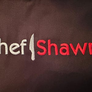 May include: A dark brown apron featuring the embroidered words "Chef Shawn." The word "Chef" is in white, the knife is silver, and "Shawn" is in red. The apron is made of a robust fabric, suitable for kitchen use.