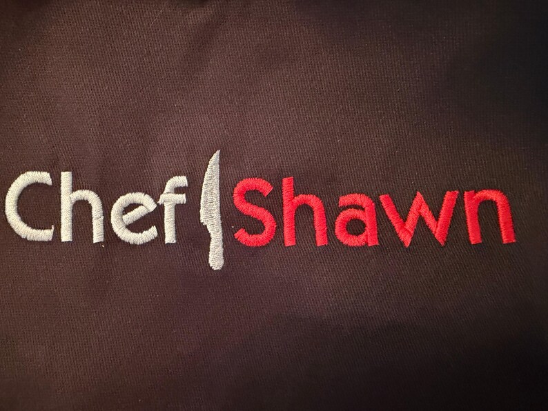 May include: A dark brown apron featuring the embroidered words "Chef Shawn." The word "Chef" is in white, the knife is silver, and "Shawn" is in red. The apron is made of a robust fabric, suitable for kitchen use.
