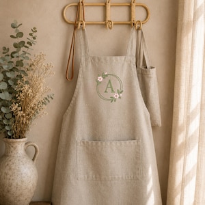 May include: A beige linen apron featuring a central embroidered design of a green circular wreath with pink flowers and the letter A in the middle. The apron has a large front pocket and is hanging from a light brown wooden wall hook.