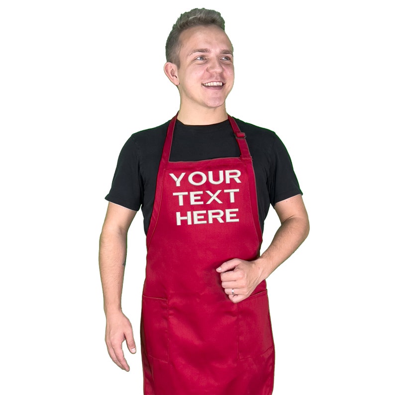 Może przedstawiać: Czerwony fartuch z białym napisem "YOUR TEXT HERE". Fartuch ma dwie przednie kieszenie i pasek na szyję. Osoba na zdjęciu ma na sobie czarny t-shirt.