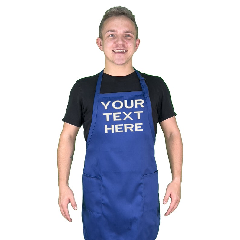 Może przedstawiać: Kr&oacute;lewski niebieski fartuch z dwiema przednimi kieszeniami i białym napisem "YOUR TEXT HERE". Fartuch jest noszony na czarnym t-shircie. Obraz jest na białym tle.