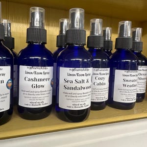 May include: A shelf of blue glass spray bottles with white labels. The labels have the text "myflowerchildo Linen/Room Spray" and different scents, including "Cashmere Glow", "Sea Salt & Sandalwood", "Cozy Cabin", "Sweater Weather", and "Ocean Shores".
