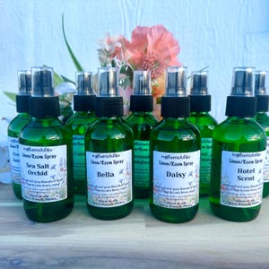 May include: A collection of green glass spray bottles with black pump nozzles, each featuring a white label with the text myflowerchildco Linen/Room Spray and specific scent names like Sea Salt Orchid, Bella, Daisy, and Hotel Scent.