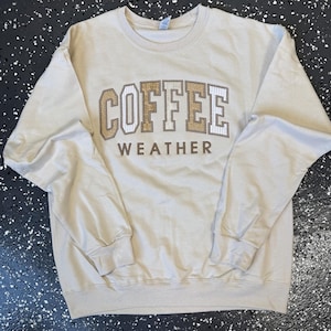 May include: A beige sweatshirt with the words "COFFEE WEATHER" in a brown, white, and tan collegiate-style design. The sweatshirt has a crew neck and long sleeves. The background is a speckled black and white surface.