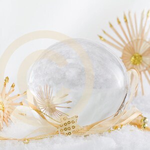 Könnte beinhalten: Eine klare Glaskugel-Dekoration mit einem weißen, schneebedeckten Inneren. Die Kugel ist von zwei beigefarbenen, sternförmigen Dekorationen mit goldenen Akzenten umgeben. Die Dekorationen befinden sich auf einer weißen, schneebedeckten Oberfläche.