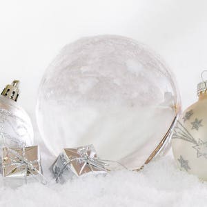 Könnte beinhalten: Drei silberne Weihnachtskugeln, darunter eine große, klare Glaskugel, eine kleinere silberne Kugel mit glitzerndem Finish und eine kleinere weiße Kugel mit silbernen Sternen. Die Kugeln liegen auf einer weißen Oberfläche, die wie Schnee aussieht.