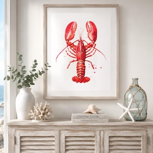 May include: A framed watercolor print of a bright red lobster centered on a white background. The artwork features detailed brushstrokes and paint splatters, displayed in a light-colored wooden frame hanging on a wall above a wooden cabinet.