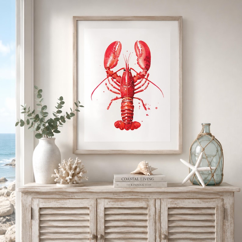 May include: A framed watercolor print of a bright red lobster centered on a white background. The artwork features detailed brushstrokes and paint splatters, displayed in a light-colored wooden frame hanging on a wall above a wooden cabinet.