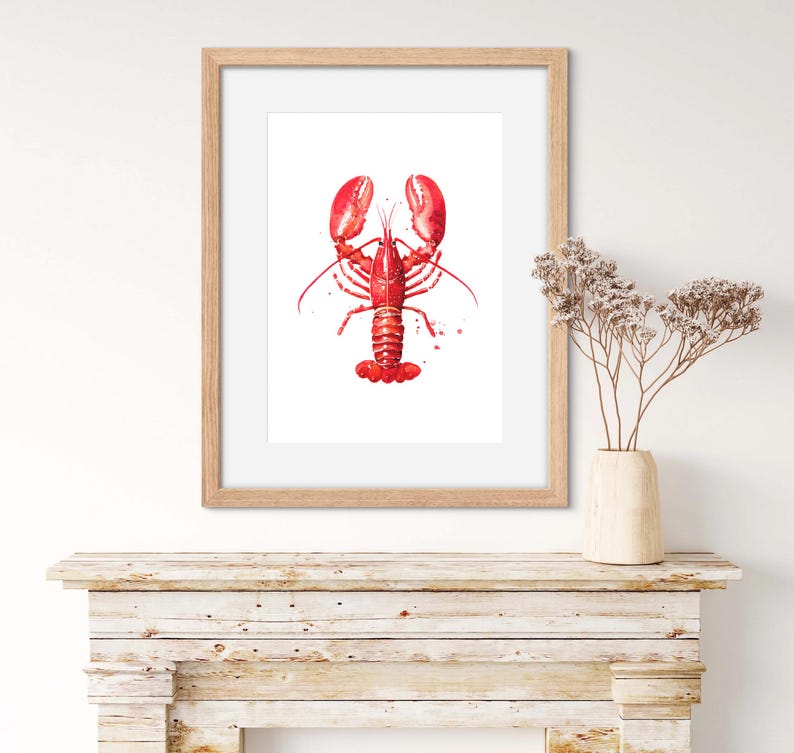May include: A framed watercolor print featuring a vibrant red lobster centered on a white background. The artwork is encased in a light-toned wooden frame, showcasing detailed brushstrokes and paint splatters that define the crustacean's form.