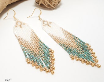 Op de afbeelding: Een paar lange, geperde oorbellen met een wit, goud en turquoise kleurenschema. De oorbellen zijn gemaakt van kleine, ronde kralen en hebben een franje-achtig ontwerp.
