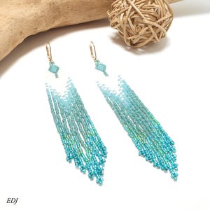 May include: A pair of long, beaded earrings with a gradient of blue and turquoise colors. The earrings have gold-colored hooks and a geometric design at the top. The beads cascade down in a fringe style, creating a flowing effect. The letters "EDJ" are visible in the bottom left corner.